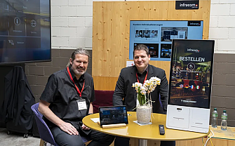 Two men sit at a table with technology and plants.