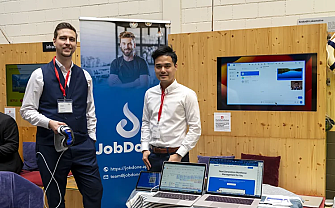 Two men stand at a trade fair booth with laptops and a presentation.