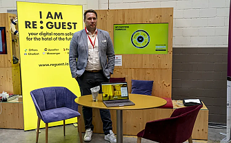 Man stands in front of a trade fair booth with laptop and advertising banner.